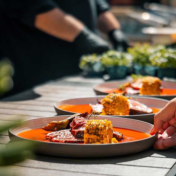Das Bild zeigt ein sorgfältig angerichtetes Gericht mit gegrilltem Fleisch, Mais und einer orangenen Sauce auf mehreren Tellern, die von einem im Hintergrund unscharf dargestellten Koch vorbereitet wurden.